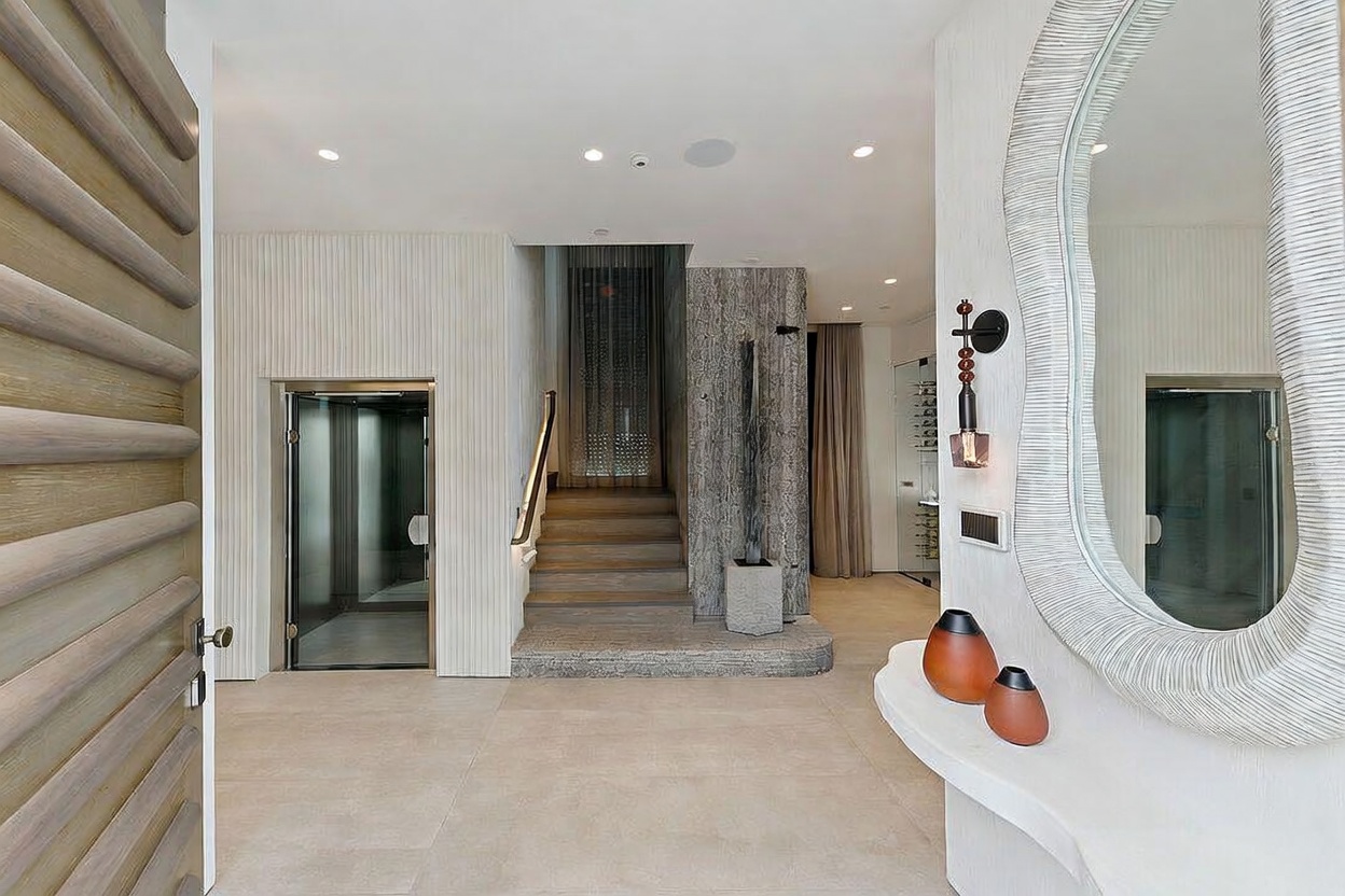 This sophisticated entryway features a modern glass-enclosed elevator and a central staircase with a stone-clad accent wall. A sculptural mirror hangs above a curved console table adorned with decorative vases, creating a welcoming and artistic focal point. The space is characterized by clean lines, neutral tones, and recessed lighting, offering a luxurious and contemporary aesthetic.