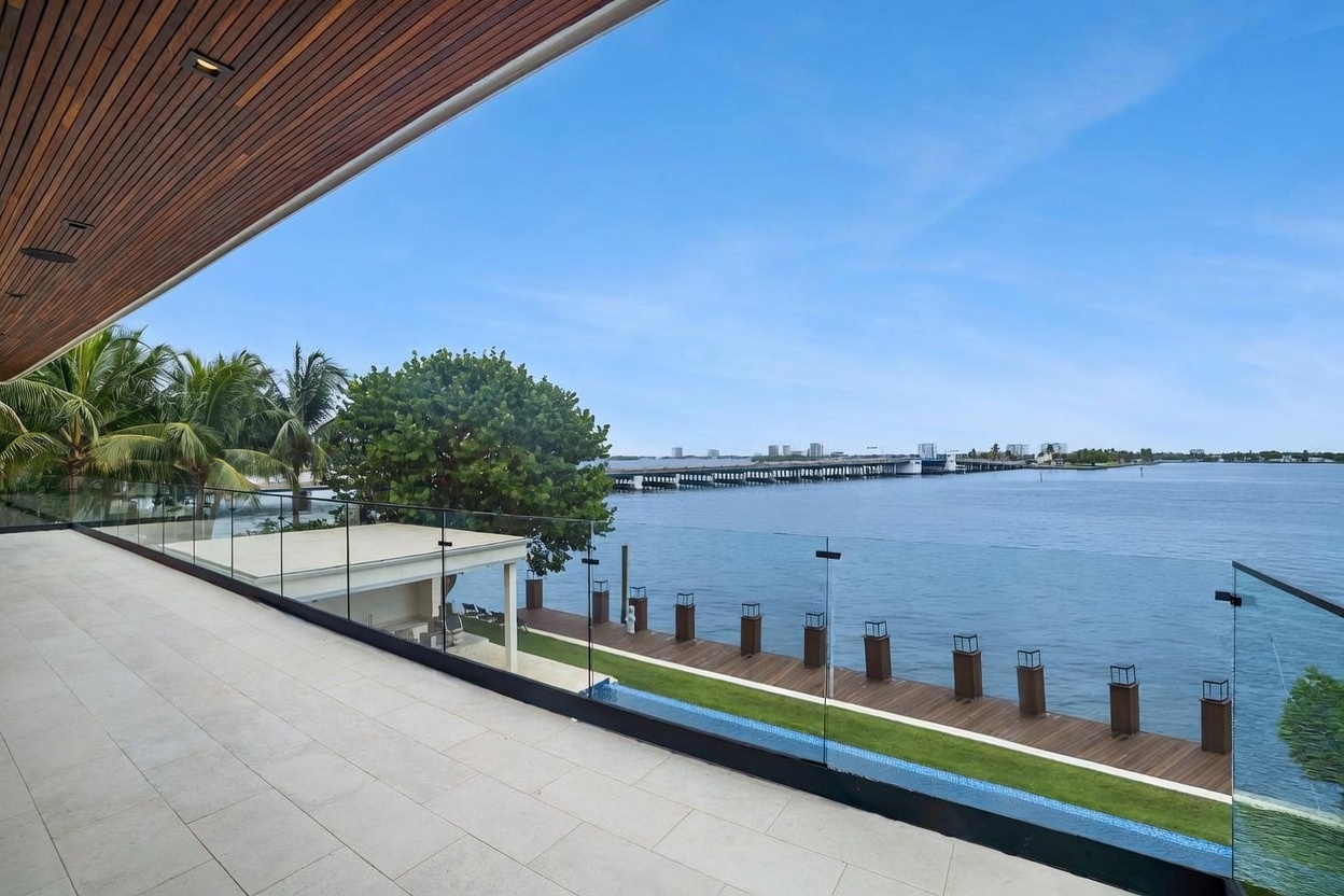 This expansive, modern balcony features light-toned stone flooring and a sleek glass railing that provides an unobstructed view of the waterfront. Below, a wooden dock extends into the water, accompanied by a small pool and a manicured lawn area. The scene is framed by a wood-paneled ceiling overhang, creating a sophisticated outdoor living space overlooking a bridge and distant cityscape.