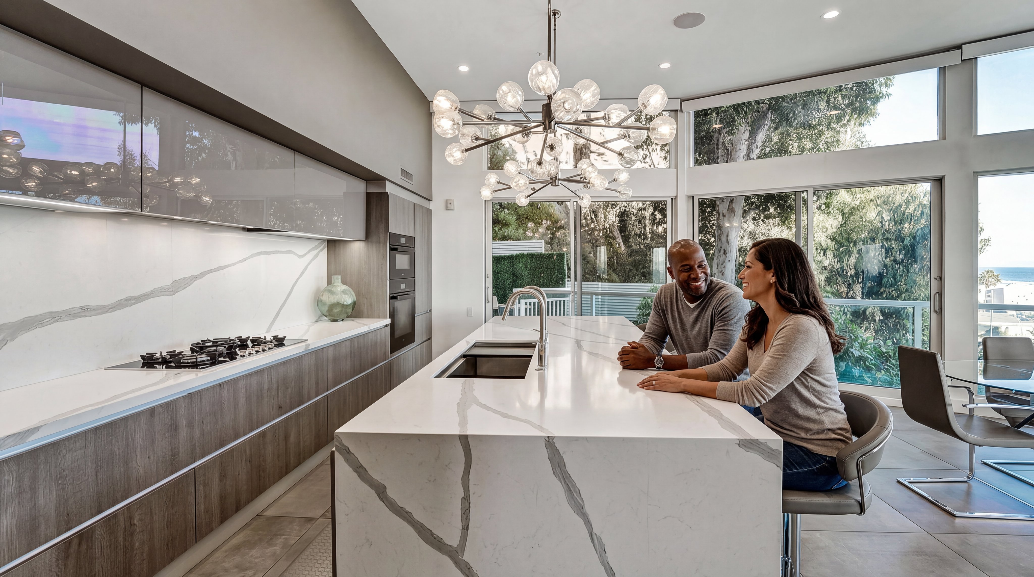 This sleek, modern kitchen features a spacious waterfall-edge marble island, warm wood cabinetry, and high-end built-in appliances. The room is flooded with natural light from floor-to-ceiling windows and accented with a striking geometric chandelier. A couple is shown seated at the island, highlighting the functional and social nature of this luxury culinary space.