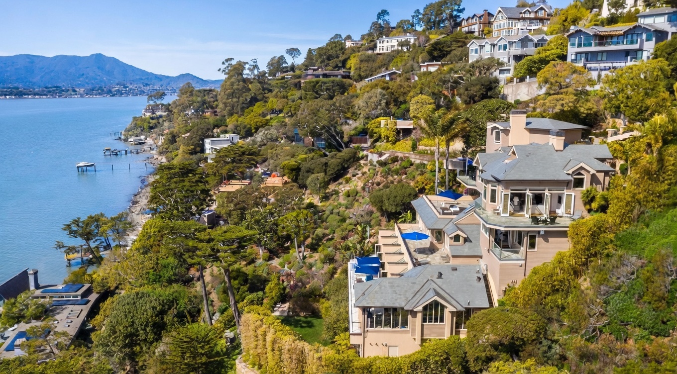 This high-angle aerial view captures a multi-level hillside residence nestled among lush greenery overlooking a tranquil bay. The home features a series of tiered decks with blue umbrellas, large windows, and a shingled roof, all integrated into the steep, landscaped terrain. In the distance, a calm body of water stretches toward rolling mountains, creating a serene and picturesque coastal setting.