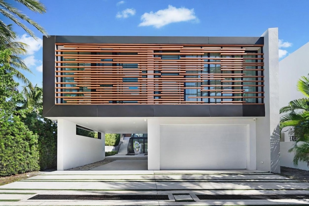 This modern, two-story residence features a striking facade characterized by a cantilevered upper level adorned with horizontal wood slats. The ground floor includes a white garage door and an open-air carport area that leads to a recessed entryway with a sculptural art piece. The driveway is composed of concrete pavers with integrated grass strips, framed by lush tropical landscaping and palm trees under a bright blue sky.