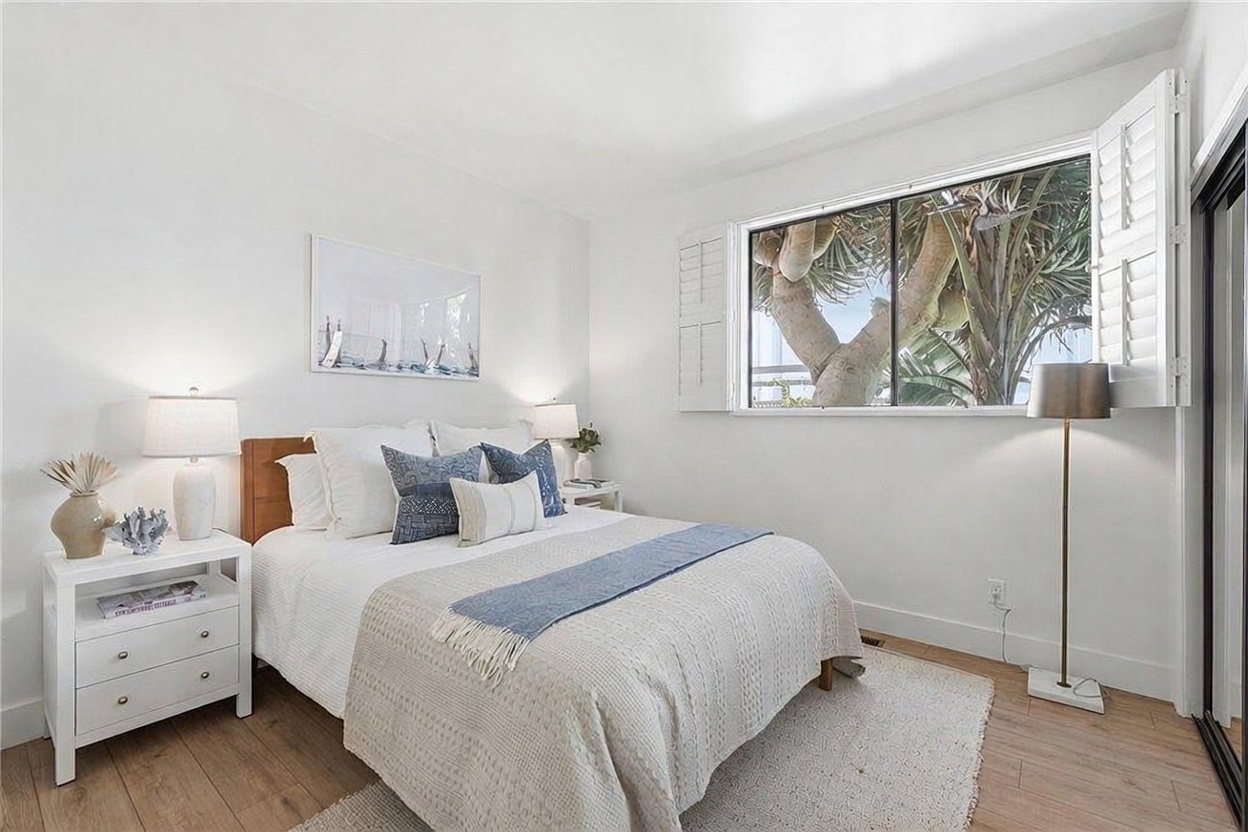 This bright and airy bedroom features light wood-toned flooring and crisp white walls, creating a clean, coastal-inspired aesthetic. The room is furnished with a queen-sized bed dressed in neutral linens and blue accent pillows, flanked by a white nightstand and a modern floor lamp. A large window with white plantation shutters allows natural light to filter in, offering a view of lush greenery outside.