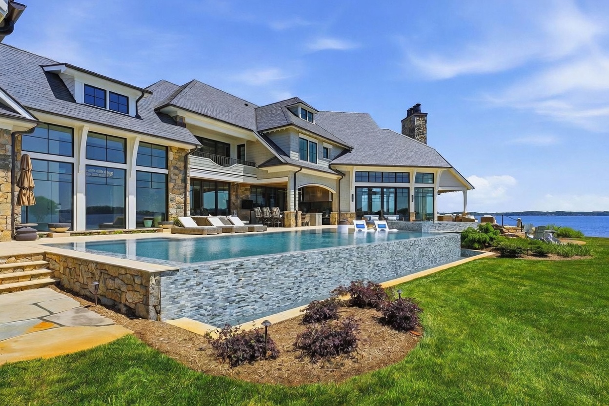 This stunning rear view of a luxury waterfront estate showcases a sprawling infinity-edge pool with a decorative tiled exterior wall. The home features a sophisticated blend of stone and siding, with large windows and a covered patio area perfect for outdoor entertaining. The manicured lawn and serene lake backdrop create an atmosphere of high-end relaxation and architectural elegance.