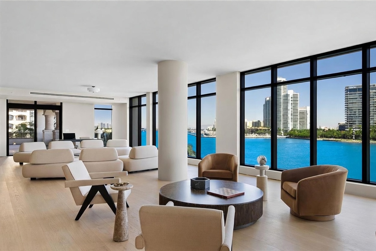 This expansive, light-filled living room features a minimalist aesthetic with light wood flooring and a neutral color palette. The space is defined by floor-to-ceiling windows that provide panoramic views of the bay and city skyline, creating a seamless connection between the interior and the water. A circular coffee table serves as a focal point, surrounded by modern, low-profile seating arrangements that emphasize the open-concept layout.