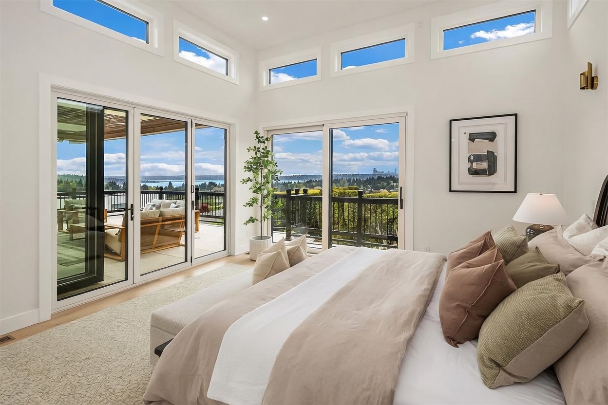 This bright and airy primary bedroom features a neutral color palette and a large bed adorned with layered pillows and a soft throw. The room is defined by expansive glass doors and tall windows that provide abundant natural light and panoramic views of the surrounding landscape and water. A minimalist framed artwork hangs on the wall, and a potted plant adds a touch of greenery to the serene, modern space.