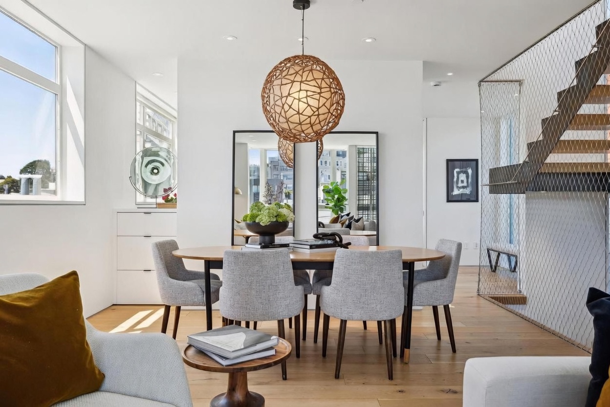 This bright and modern dining area features a round wooden table surrounded by four upholstered gray chairs, centered beneath a large, textured spherical pendant light. The space is anchored by two tall, black-framed mirrors that reflect the room, adding depth and a sense of openness. To the right, a staircase with a unique metal mesh railing leads upward, while light wood flooring runs throughout the open-concept living space.