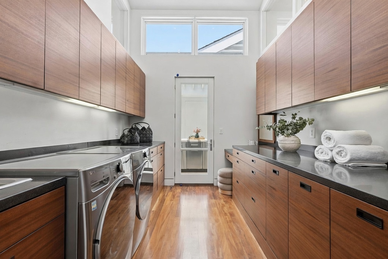 This modern laundry room features sleek, wood-toned cabinetry providing ample storage space above and below the dark countertops. A pair of stainless steel laundry appliances is positioned on the left, while a built-in desk area with a stool sits on the right. The room is brightened by a transom window above a glass-paneled door, and the space is finished with warm-toned wood flooring.