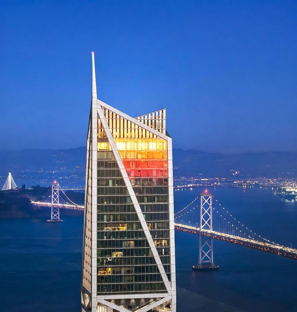 This striking aerial perspective captures a modern, angular skyscraper at twilight, featuring a distinctive spire and a facade of glass and metal. The building is positioned prominently against a backdrop of the San Francisco Bay, with the iconic Bay Bridge illuminated in the distance. The composition emphasizes the tower's unique architectural geometry and its commanding presence within the urban waterfront landscape.