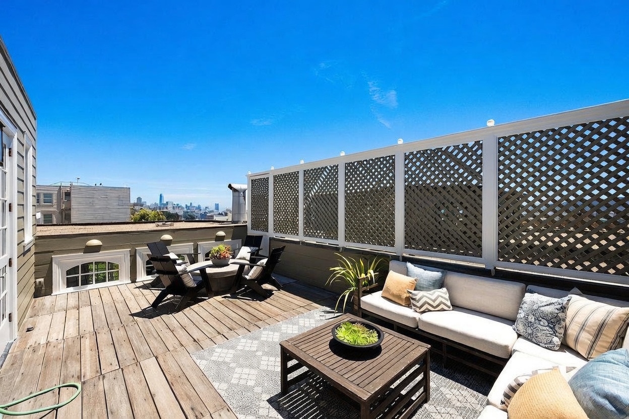 This spacious rooftop deck features weathered wood decking and a comfortable outdoor seating area with a sectional sofa and a central coffee table. A lattice privacy screen runs along the perimeter, while a small dining set sits in the corner, all set against a clear blue sky with a distant city skyline view. The space offers an inviting, open-air atmosphere perfect for entertaining or relaxing outdoors.