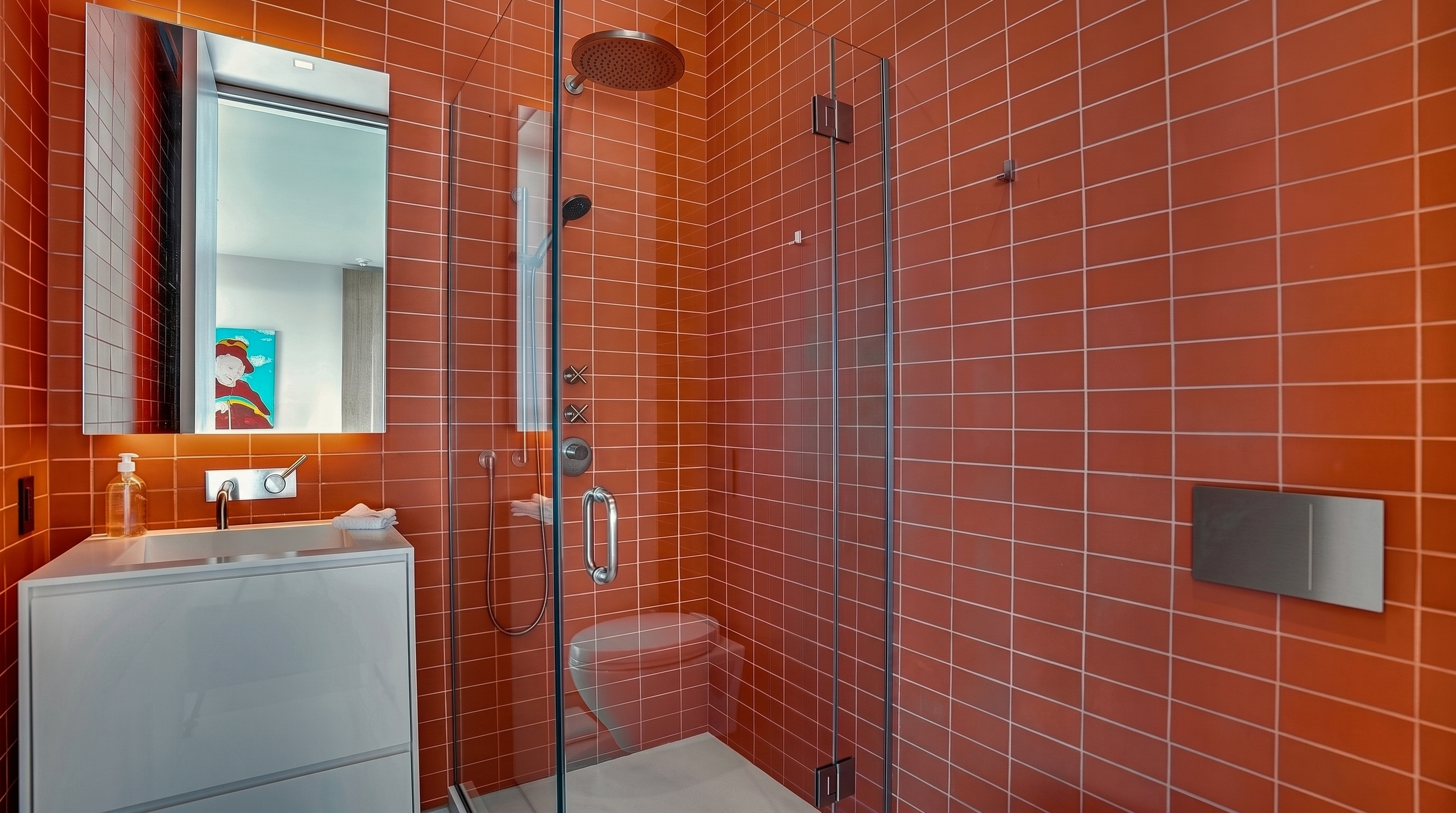 This modern bathroom features vibrant, coral-colored rectangular wall tiles that create a bold and energetic aesthetic. A sleek white vanity with a minimalist faucet sits beneath a rectangular mirror, while a glass-enclosed walk-in shower occupies the corner. The space is efficiently designed with a wall-mounted toilet and polished chrome fixtures, offering a contemporary and clean look.