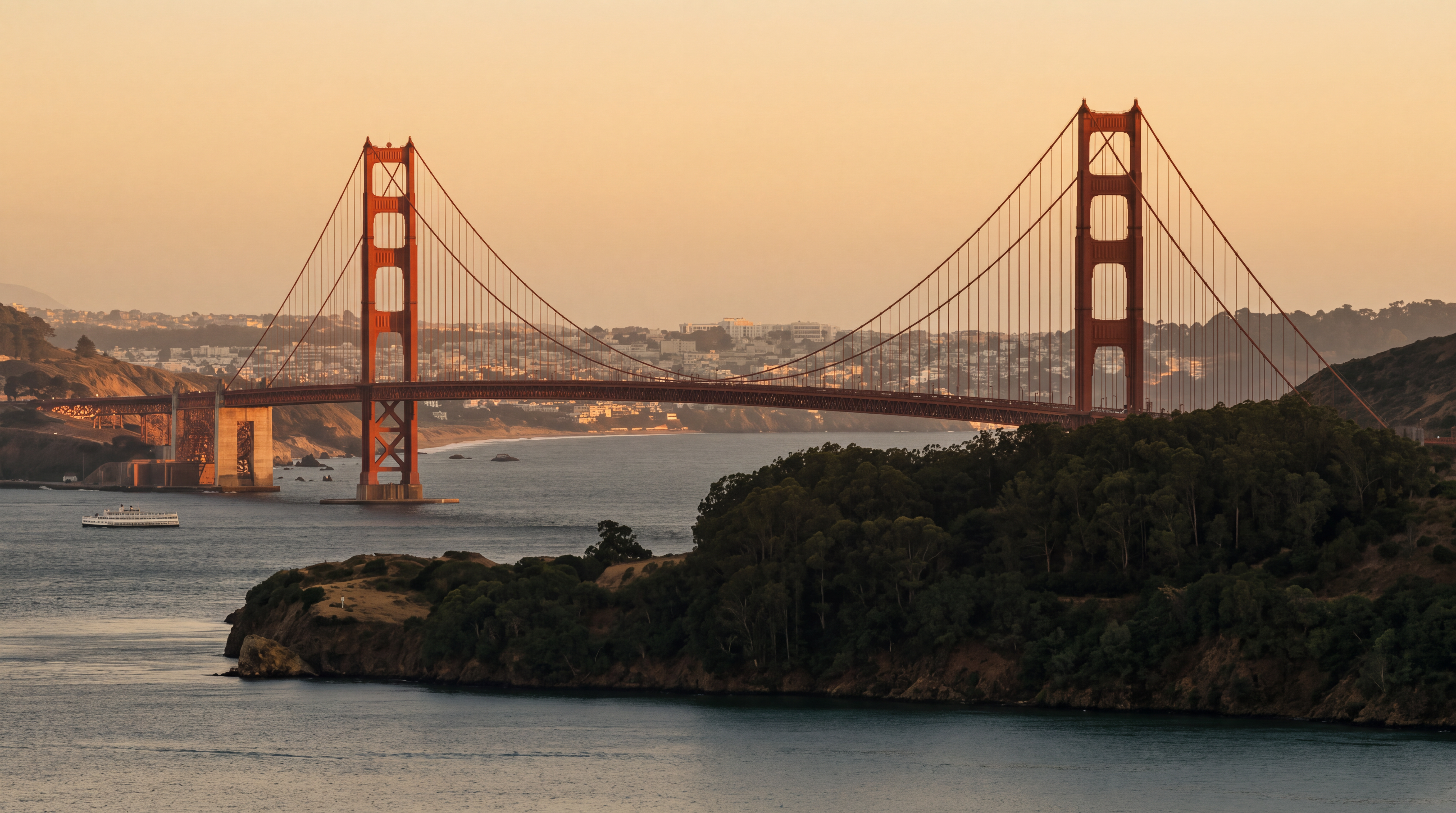 This scenic, wide-angle shot captures the iconic Golden Gate Bridge at sunset, showcasing its distinctive orange towers against a soft, gradient sky. The perspective is elevated, looking across the calm waters of the bay toward the bridge and the distant city skyline. The foreground features a lush, dark green headland that provides a natural frame for the bridge, emphasizing the property's proximity to world-famous landmarks.