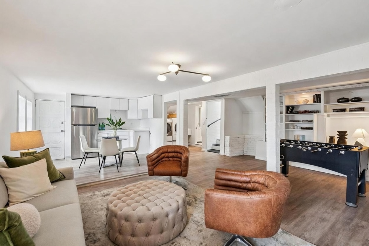 This open-concept living area features a modern, neutral aesthetic with light wood-look flooring and white-painted brick accents. The space includes a comfortable seating area with a tufted ottoman and leather swivel chairs, transitioning into a dining nook and a game area equipped with a foosball table. The bright, airy layout is enhanced by a contemporary multi-arm light fixture and a seamless flow between the living, dining, and recreational zones.