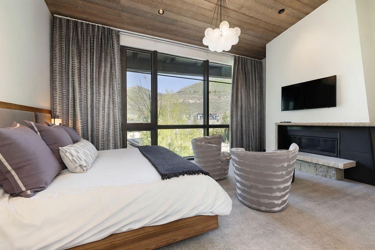 This primary bedroom features a contemporary design with a wood-paneled ceiling and large windows that frame a scenic mountain view. The room is furnished with a plush bed, two patterned armchairs, and a fireplace set into a white wall with a stone hearth. The neutral color palette and soft textures create a serene and luxurious atmosphere.