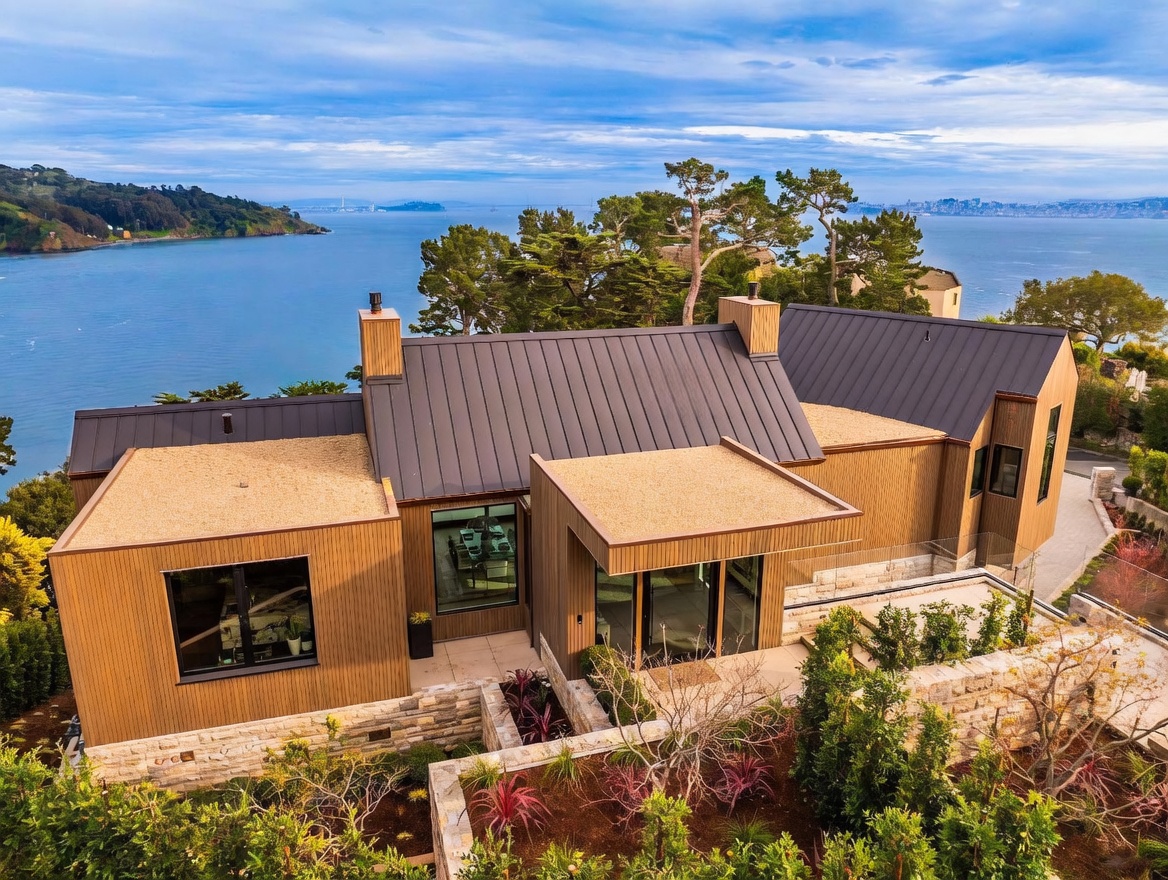 This elevated aerial perspective captures a modern, multi-gabled residence set against a stunning coastal backdrop. The home features vertical wood-toned siding, dark metal roofing, and a combination of flat-roofed sections with gravel surfaces, all integrated into a tiered landscape with stone retaining walls. The expansive view includes a calm bay, distant hills, and a glimpse of a bridge, emphasizing the property's serene and scenic location.