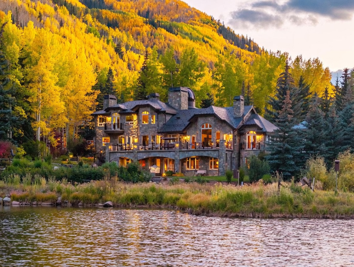 This grand, multi-story stone estate is captured from across a tranquil pond, showcasing its impressive architectural scale and warm, inviting glow at dusk. The residence features a complex roofline with multiple gables, a prominent stone chimney, and expansive decks that overlook the water and the vibrant, golden-hued autumn forest backdrop. The composition emphasizes the home's integration into the natural landscape, highlighting its luxurious mountain-style aesthetic.