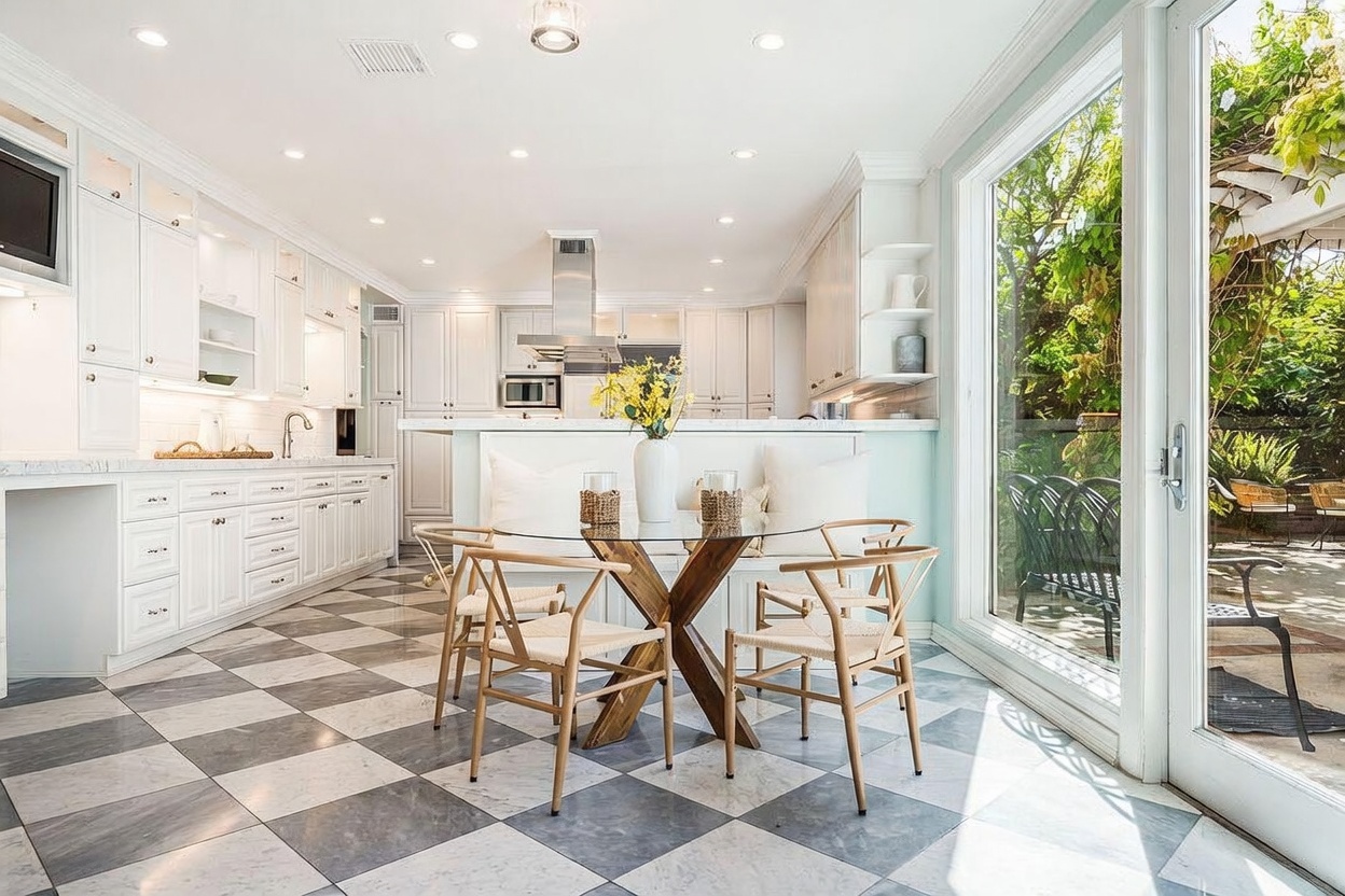 This bright, airy kitchen features a classic checkerboard stone floor and crisp white cabinetry, creating a clean and timeless aesthetic. A central dining area includes a glass-topped table with light wood chairs, positioned against a built-in bench seat that overlooks a lush outdoor garden through large glass doors. The space is illuminated by recessed lighting and offers a seamless transition between indoor cooking areas and the natural light streaming in from the patio.
