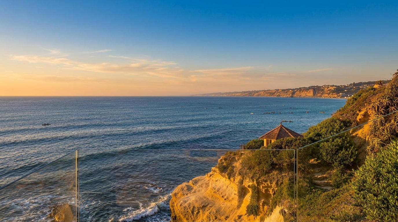 This image captures a breathtaking oceanfront view from a glass-railed balcony, showcasing a rugged cliffside landscape meeting the deep blue sea. A small, charming gazebo with a shingled roof sits nestled among the coastal greenery on the cliff edge, providing a serene focal point. The perspective offers a cinematic, wide-angle look at the expansive coastline, emphasizing the property's prime location and natural beauty.