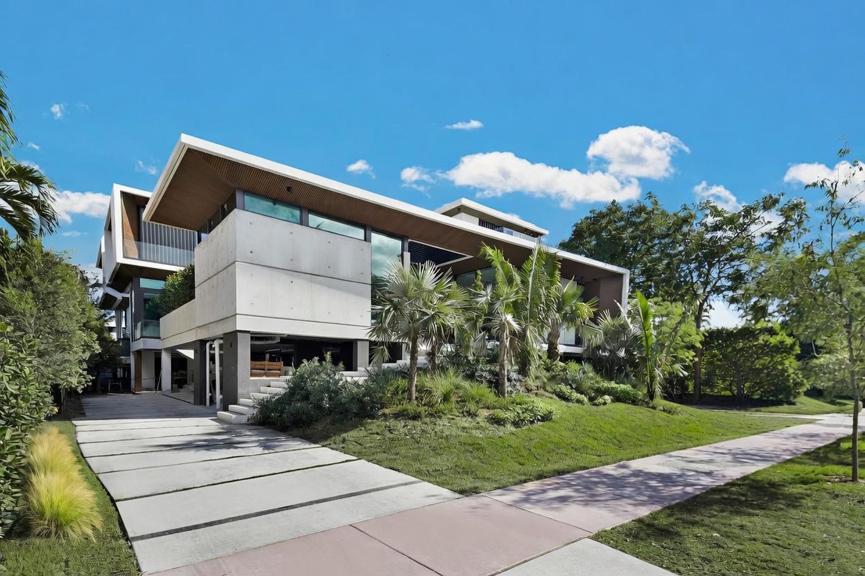 This modern architectural residence features a striking design with exposed concrete walls, clean lines, and a prominent cantilevered roofline. The home is elevated above a paved driveway and is surrounded by lush, manicured landscaping with palm trees and native shrubs. The perspective captures the home from a low angle along the sidewalk, emphasizing its contemporary aesthetic and integration with the natural environment.