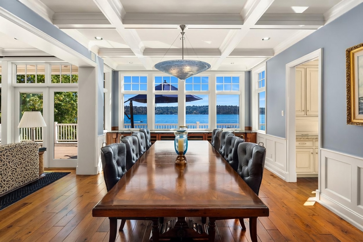 This elegant dining room features a long, polished wooden table surrounded by tufted black leather chairs, set against a backdrop of expansive windows overlooking a serene lake. The room is defined by a sophisticated coffered ceiling, light blue walls with white wainscoting, and warm hardwood flooring. The perspective is centered and symmetrical, emphasizing the luxurious atmosphere and the seamless connection between the interior space and the scenic waterfront view.