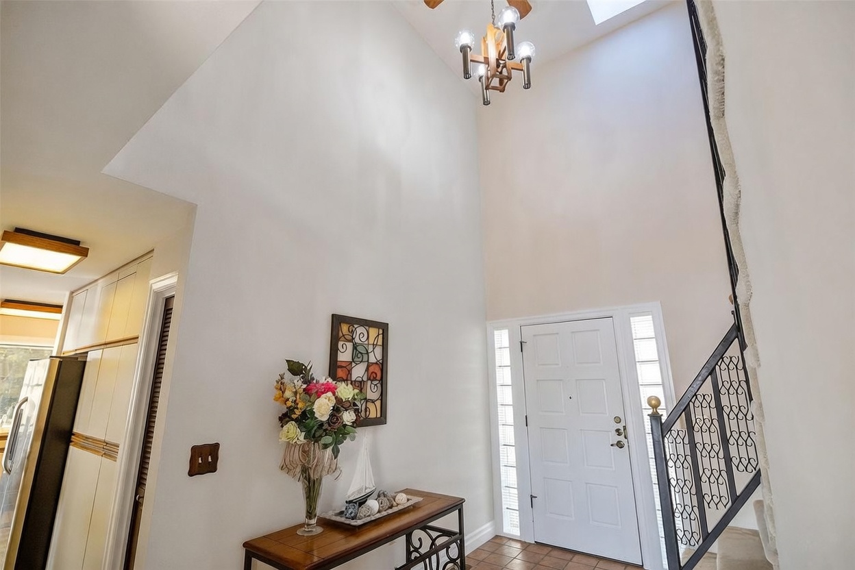 This bright and airy entryway features a dramatic double-height ceiling with a skylight and a rustic-style chandelier. A staircase with a decorative black metal railing leads to the upper level, while a console table with floral decor sits against the wall. The space is finished with light-toned walls and tiled flooring, creating a welcoming and open atmosphere.