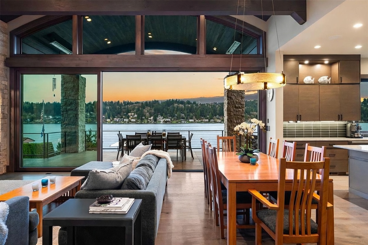 This open-concept living and dining area features a seamless transition to an outdoor deck through expansive sliding glass doors that frame a scenic lake view. The space is anchored by a comfortable gray sofa and a warm-toned wooden dining set, illuminated by a modern circular pendant light. The interior design emphasizes a connection to nature, with rich wood tones and stone accents that complement the tranquil waterfront setting.