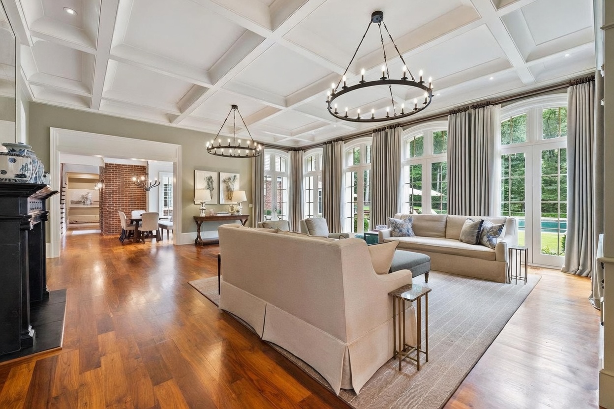 This elegant living room features a coffered ceiling, warm wood flooring, and a neutral color palette that creates a sophisticated atmosphere. Large windows with floor-to-ceiling drapery allow abundant natural light to fill the space, while two circular chandeliers provide a focal point above the seating area. A dark fireplace surround adds a striking contrast to the room, which flows seamlessly into an adjacent dining area.