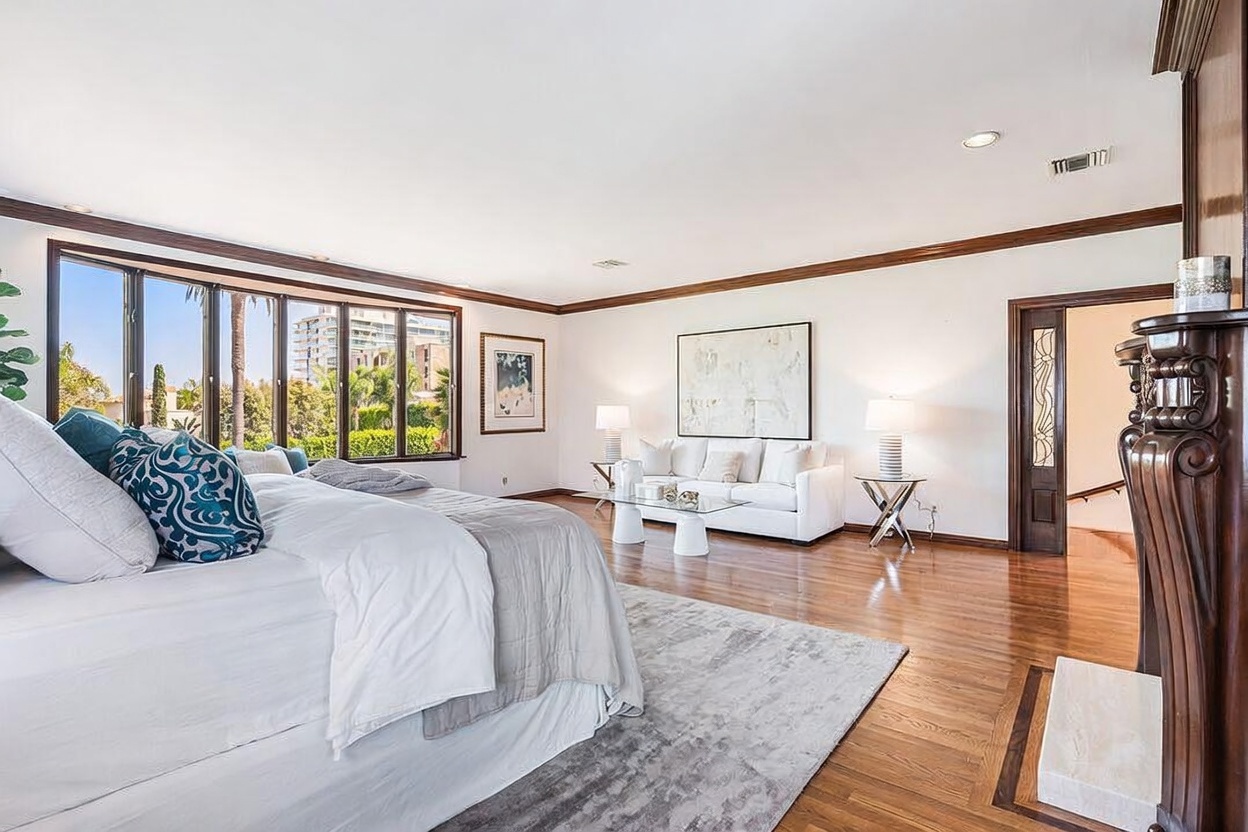 This spacious primary bedroom features polished hardwood floors and a large area rug, creating a warm and inviting atmosphere. The room includes a comfortable seating area with a white sofa and glass coffee table, positioned near large windows that offer views of the outdoors. A dark wood-framed doorway leads to an adjacent space, while a decorative fireplace surround adds a touch of traditional elegance to the room's design.