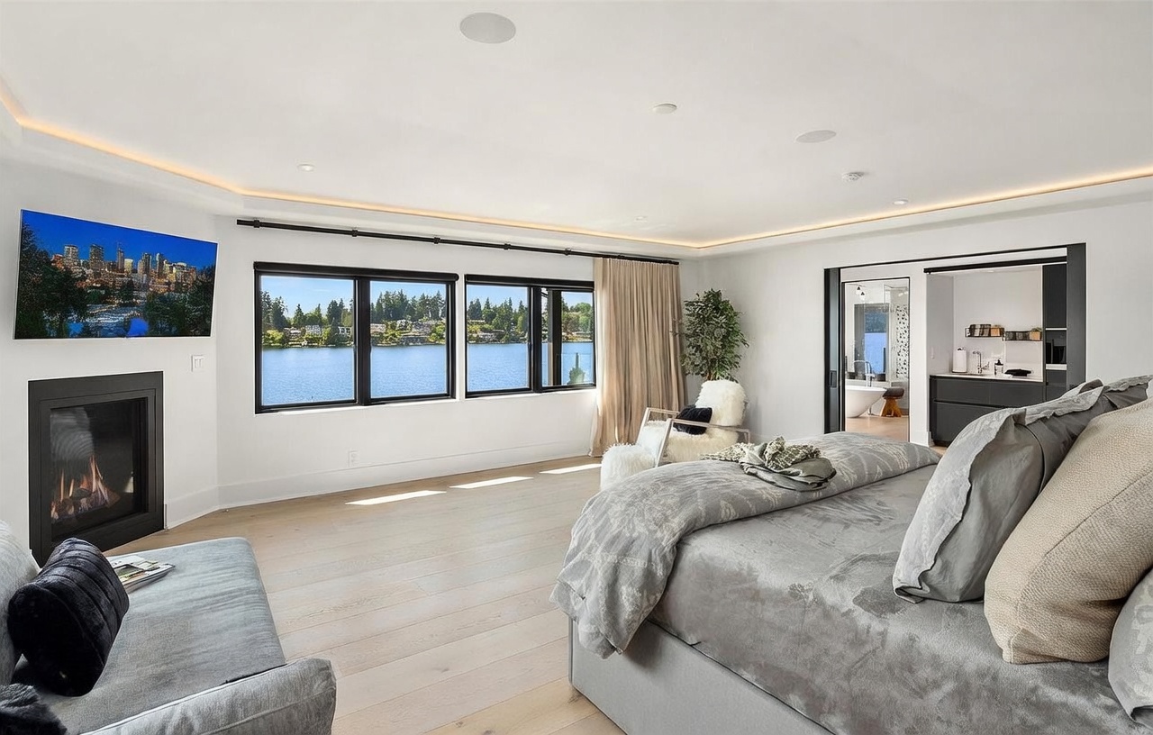 This spacious primary bedroom features light wood flooring and a neutral color palette, centered around a large bed with a textured gray duvet. A fireplace with a dark surround is positioned to the left, while a wall of large windows provides expansive views of the water and surrounding trees. To the right, a sliding door opens into an adjacent bathroom, creating an open and airy flow throughout the suite.