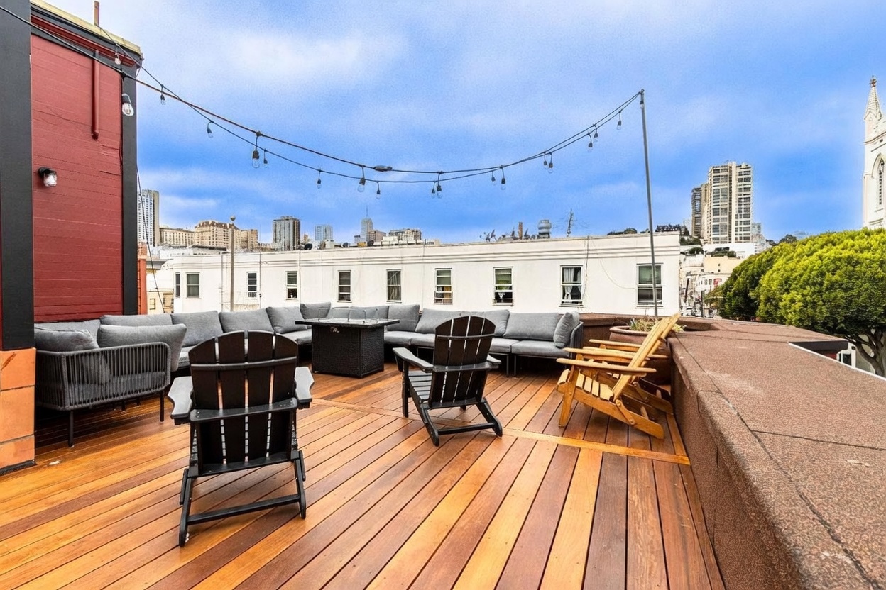 This expansive rooftop deck features warm-toned wood decking and a large, L-shaped sectional sofa arranged around a central fire pit. The space is accented by black Adirondack chairs and wooden lounge chairs, all set against a backdrop of urban city views under a cloudy sky. String lights are strung overhead, creating an inviting atmosphere for outdoor entertaining.