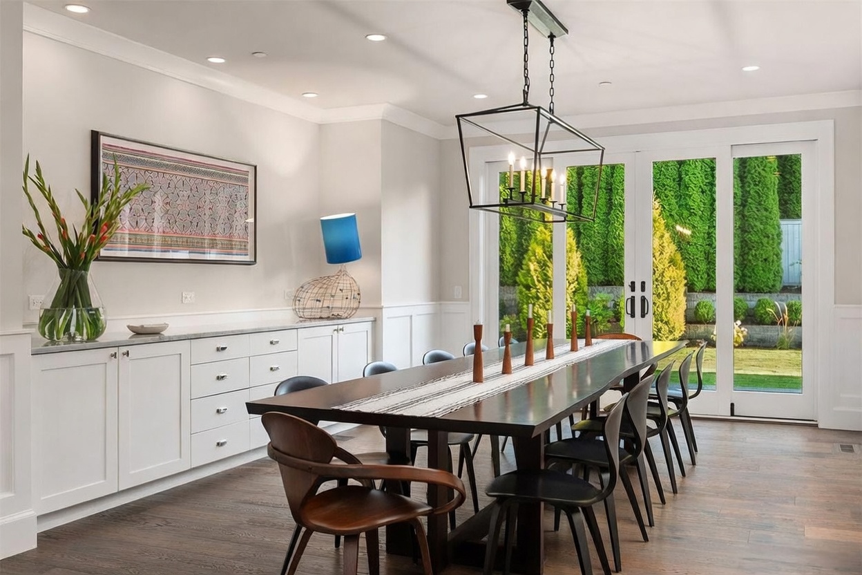 This elegant dining room features a long, dark wood table surrounded by modern chairs, positioned beneath a large, geometric black metal chandelier. To the left, a built-in white sideboard with stone countertops provides ample storage and display space, accented by a vibrant piece of wall art and a unique blue-shaded lamp. Large glass doors at the rear of the room offer a clear view of the lush green garden outside, filling the space with natural light.