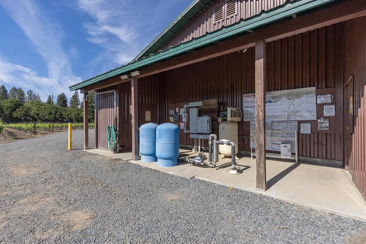 This side view of a utility building features a covered concrete porch area housing industrial water filtration equipment, including two large blue pressure tanks and a compressor. The structure is clad in dark brown corrugated metal siding and sits adjacent to a gravel path that leads toward a vineyard in the background. The scene captures a functional, agricultural-style workspace under a bright, clear sky.
