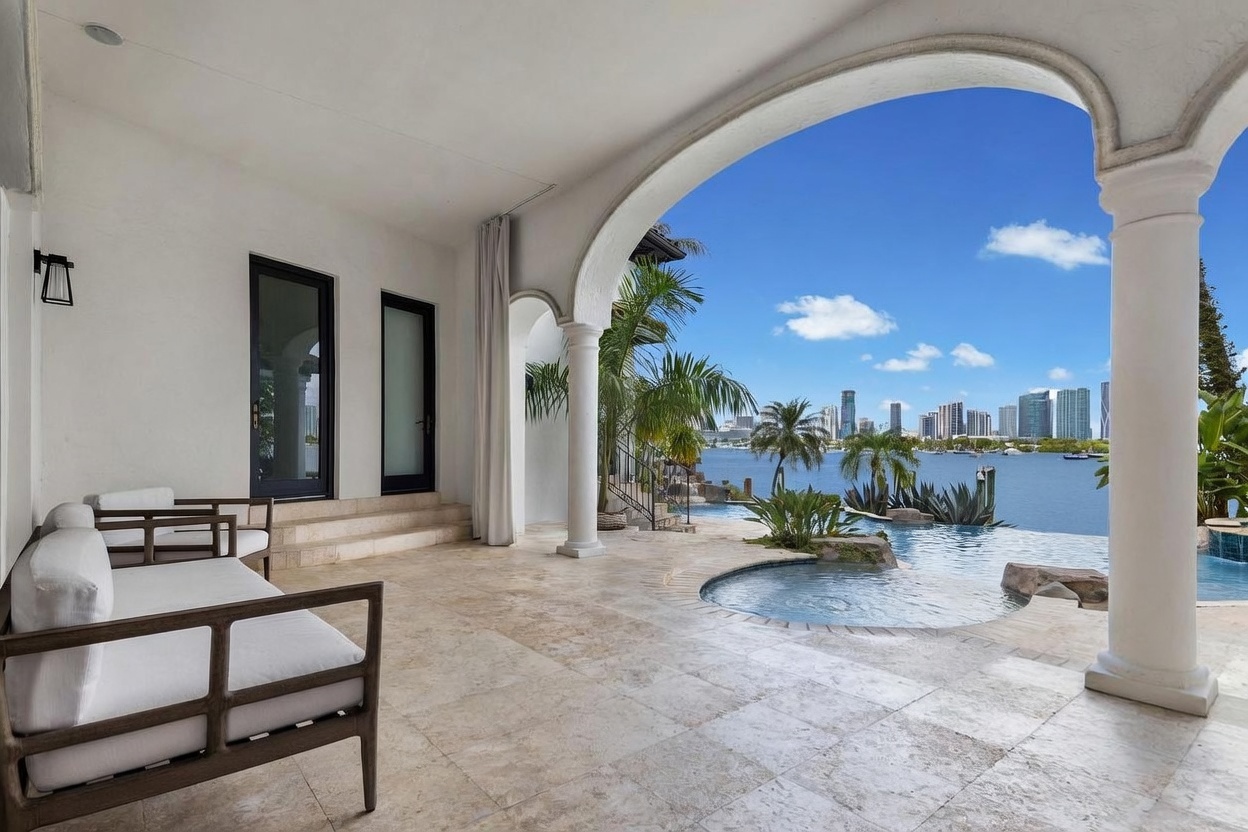This luxurious covered patio features light-toned stone flooring and elegant white columns that frame a stunning waterfront view. The space includes comfortable outdoor seating and transitions seamlessly into a private pool and spa area overlooking a distant city skyline. The open-air design creates a serene, resort-like atmosphere perfect for relaxation and entertaining.