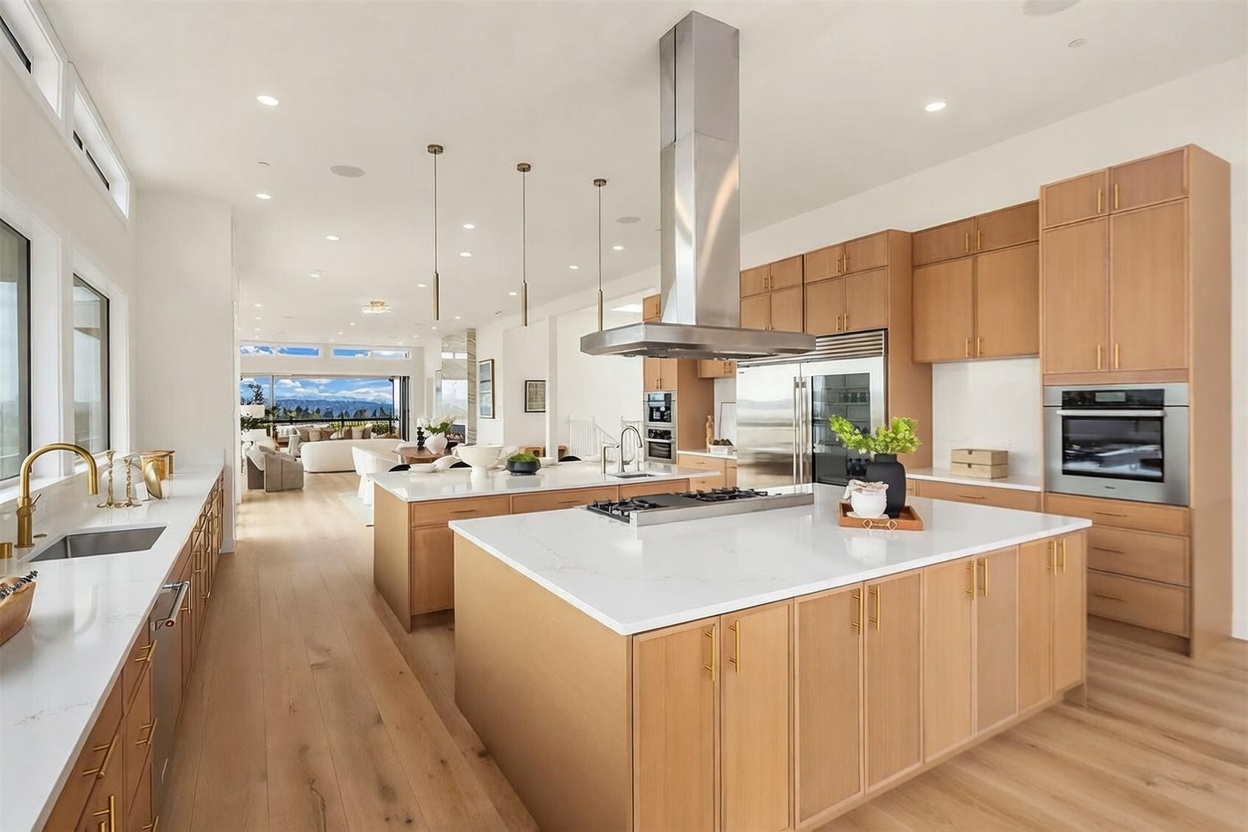 This expansive, modern kitchen features light wood cabinetry, a large central island with a gas cooktop, and a secondary island with a sink. The space is illuminated by sleek pendant lighting and recessed lights, with a stainless steel range hood serving as a focal point. The open-concept design flows seamlessly into a bright living area with large windows that offer a view of the outdoors.