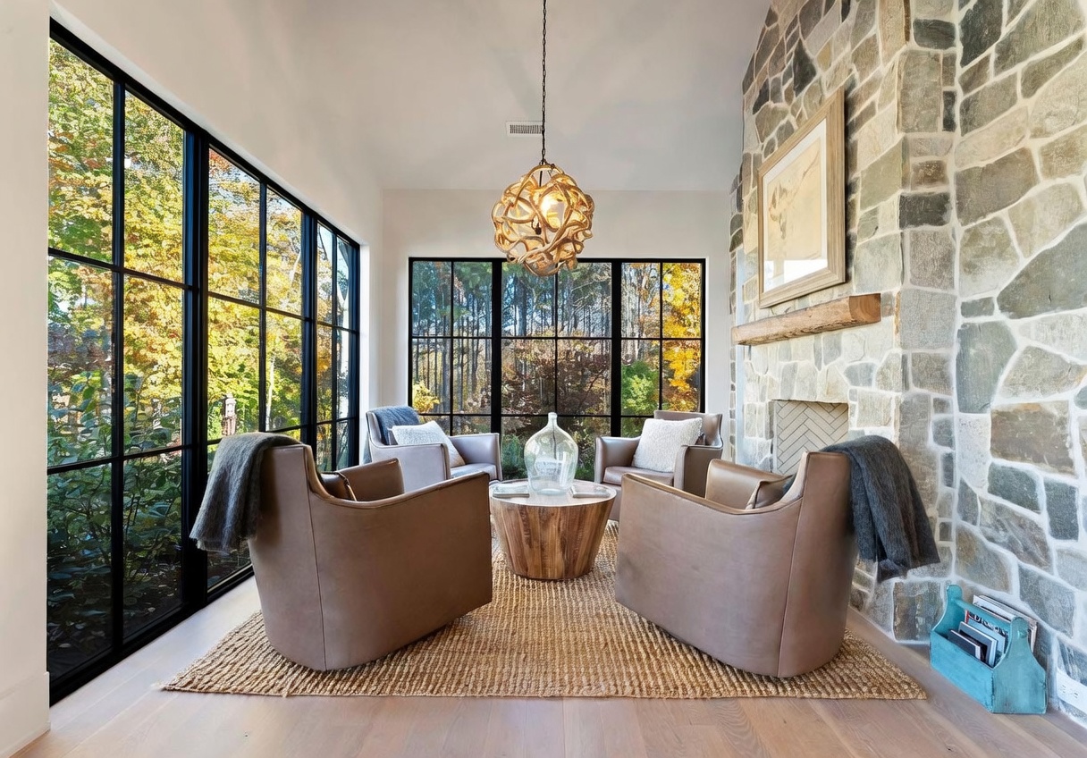 This bright sunroom features expansive windows with black frames that offer panoramic views of the surrounding trees. The space is anchored by a stone-clad chimney breast with a rustic wood mantel and is furnished with four comfortable leather armchairs arranged around a round wooden coffee table. A textured natural fiber rug sits atop light wood flooring, while a unique sculptural pendant light hangs from the ceiling, creating a warm and inviting atmosphere.