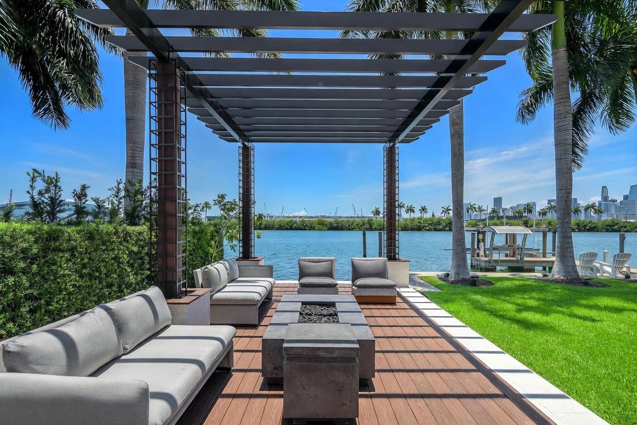 This luxurious outdoor living space features a modern pergola covering a spacious wooden deck with a central fire pit and comfortable lounge seating. The area overlooks a serene waterfront with a private dock, framed by lush green grass and tall palm trees under a clear blue sky. The scene captures a sophisticated, resort-style atmosphere perfect for waterfront entertaining.