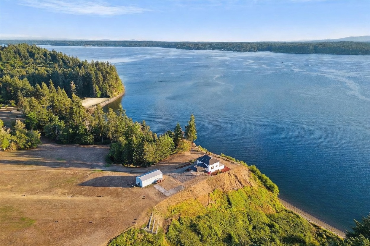 This high-angle aerial shot captures a secluded waterfront property situated on a prominent bluff overlooking a vast, calm body of water. The scene features a white house and a detached metal outbuilding set against a backdrop of dense evergreen forest and expansive, open fields. The perspective emphasizes the dramatic coastal location and the expansive natural landscape surrounding the residence.