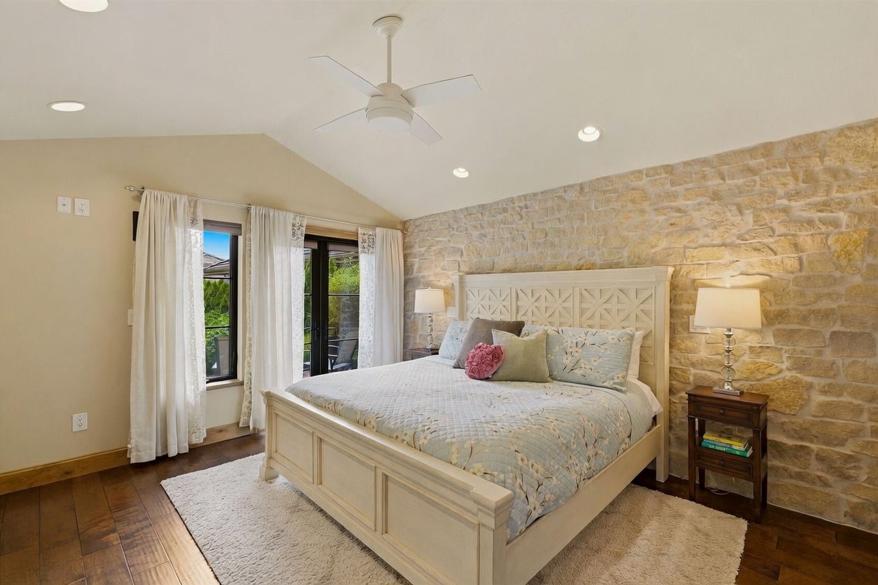 This primary bedroom features a striking stone-clad accent wall behind a large, light-colored wooden bed frame. The room is illuminated by recessed lighting and a central ceiling fan, with a plush area rug resting on dark wood flooring. Large windows draped in white curtains provide natural light and access to an outdoor space, creating a warm and inviting atmosphere.