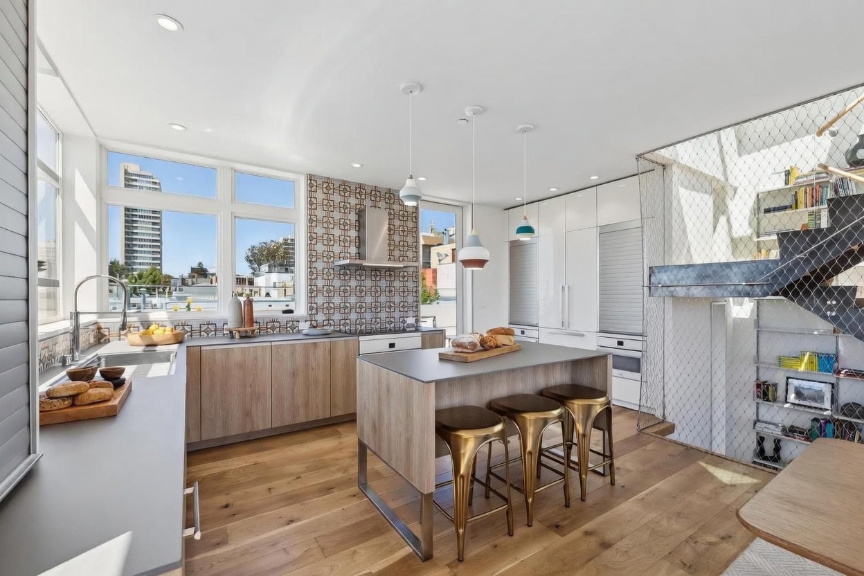 This modern, open-concept kitchen features light wood cabinetry, a central island with three metallic bar stools, and a striking patterned tile backsplash. Large windows provide ample natural light and an urban view, while a unique staircase with a wire mesh railing adds an industrial architectural element to the space. The room is accented by three pendant lights hanging over the island, creating a bright and contemporary atmosphere.
