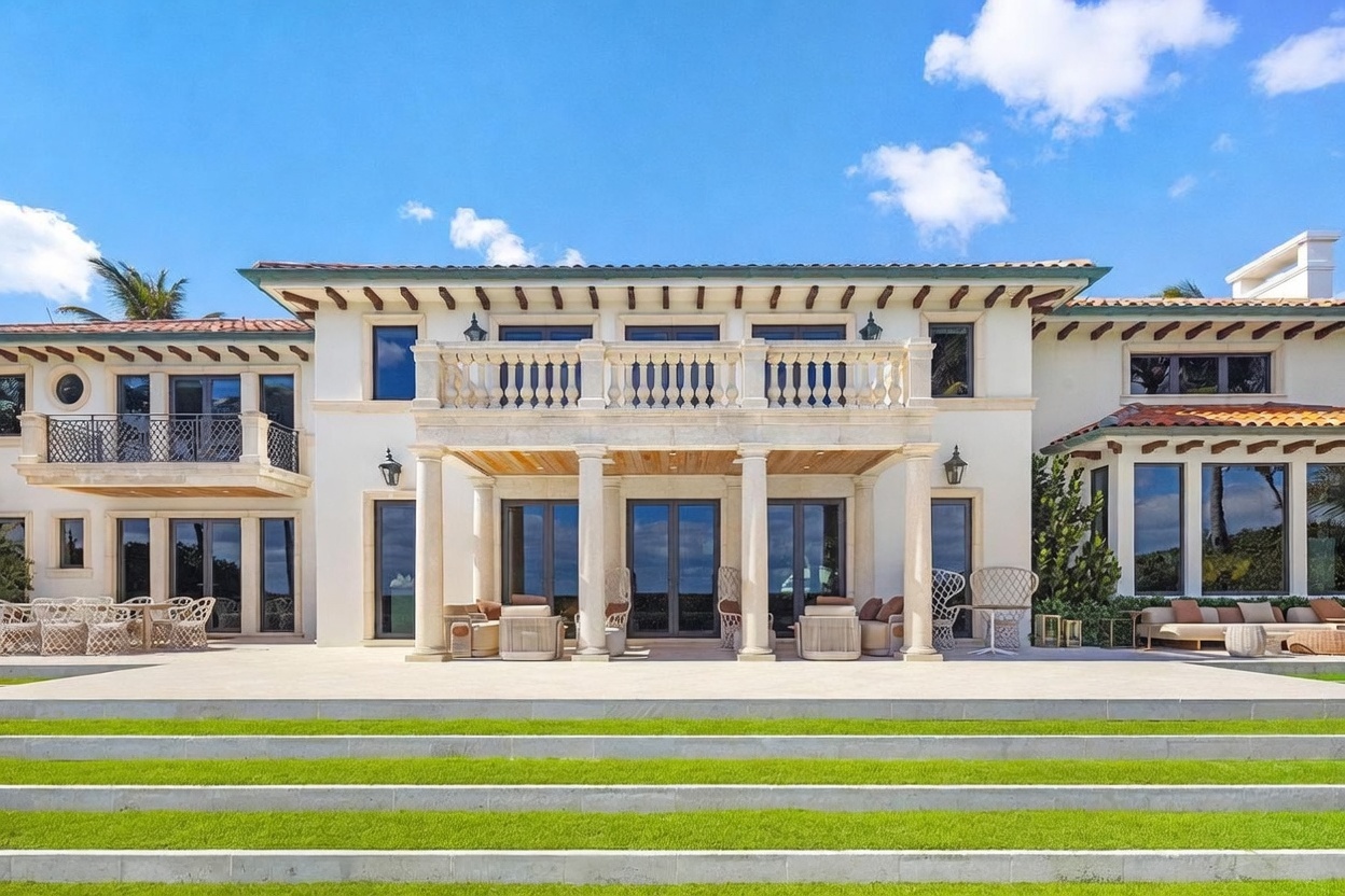 This expansive rear exterior view showcases a grand Mediterranean-style estate with a symmetrical facade, featuring a prominent stone-columned loggia and a second-story balcony with ornate balustrades. The home is characterized by its light-colored stucco walls, dark-framed windows, and a classic terracotta tile roof. A wide, paved patio area transitions into a tiered lawn, creating a sophisticated and inviting outdoor living space perfect for entertaining.