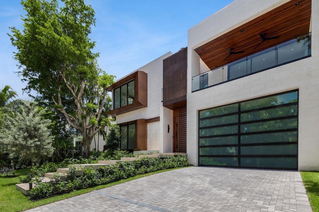 This modern luxury residence features a striking facade composed of light-toned stone panels, dark metal accents, and warm wood-paneled soffits. The front elevation includes a large glass-paneled garage door, a recessed entryway with a decorative screen, and cantilevered window boxes that add architectural depth. A paved driveway leads to the entrance, complemented by tiered landscaping and a mature tree that frames the home against a clear blue sky.