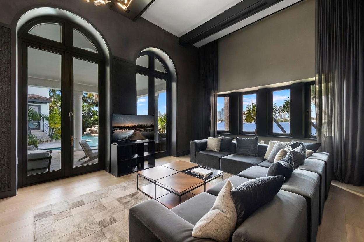 This spacious living room features a large, dark-toned sectional sofa arranged around a set of modular coffee tables on a patterned area rug. The room is defined by its dramatic dark walls, high ceilings, and expansive arched glass doors that provide a seamless view of the outdoor pool and waterfront. The combination of modern furniture and architectural elements creates a sophisticated, high-end atmosphere with abundant natural light.