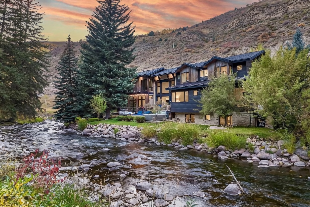 This stunning mountain residence is captured from a low-angle perspective across a flowing river, showcasing its multi-level architectural design. The home features a blend of dark siding, natural wood accents, and stone foundations, nestled against a dramatic, rugged hillside under a vibrant sunset sky. A landscaped lawn with a patio and hot tub area provides a seamless transition between the luxury living space and the natural riparian environment.