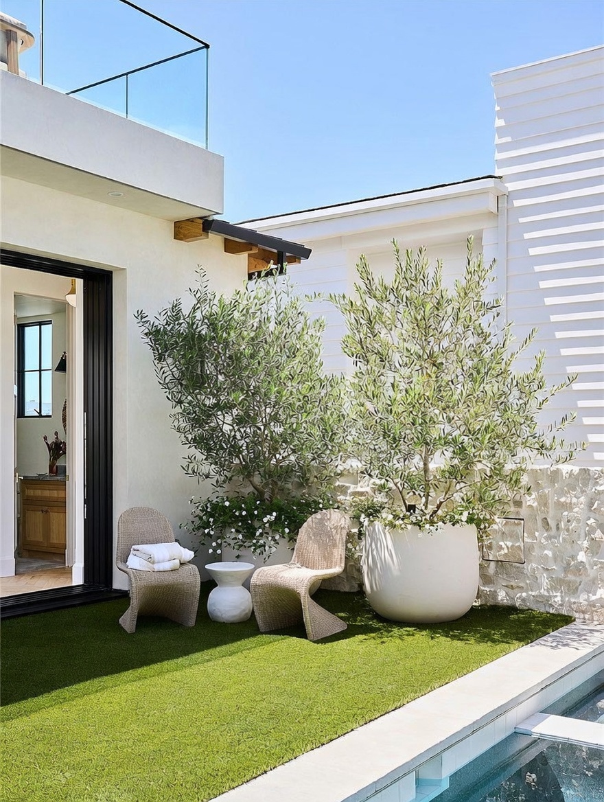 This modern outdoor patio space features a clean, minimalist aesthetic with artificial turf and a sleek swimming pool edge. Two sculptural woven chairs are positioned around a small white pedestal table, set against a backdrop of lush potted olive trees and a textured stone wall. The area seamlessly connects to the interior through a large glass sliding door, creating a bright and inviting transition between indoor and outdoor living.