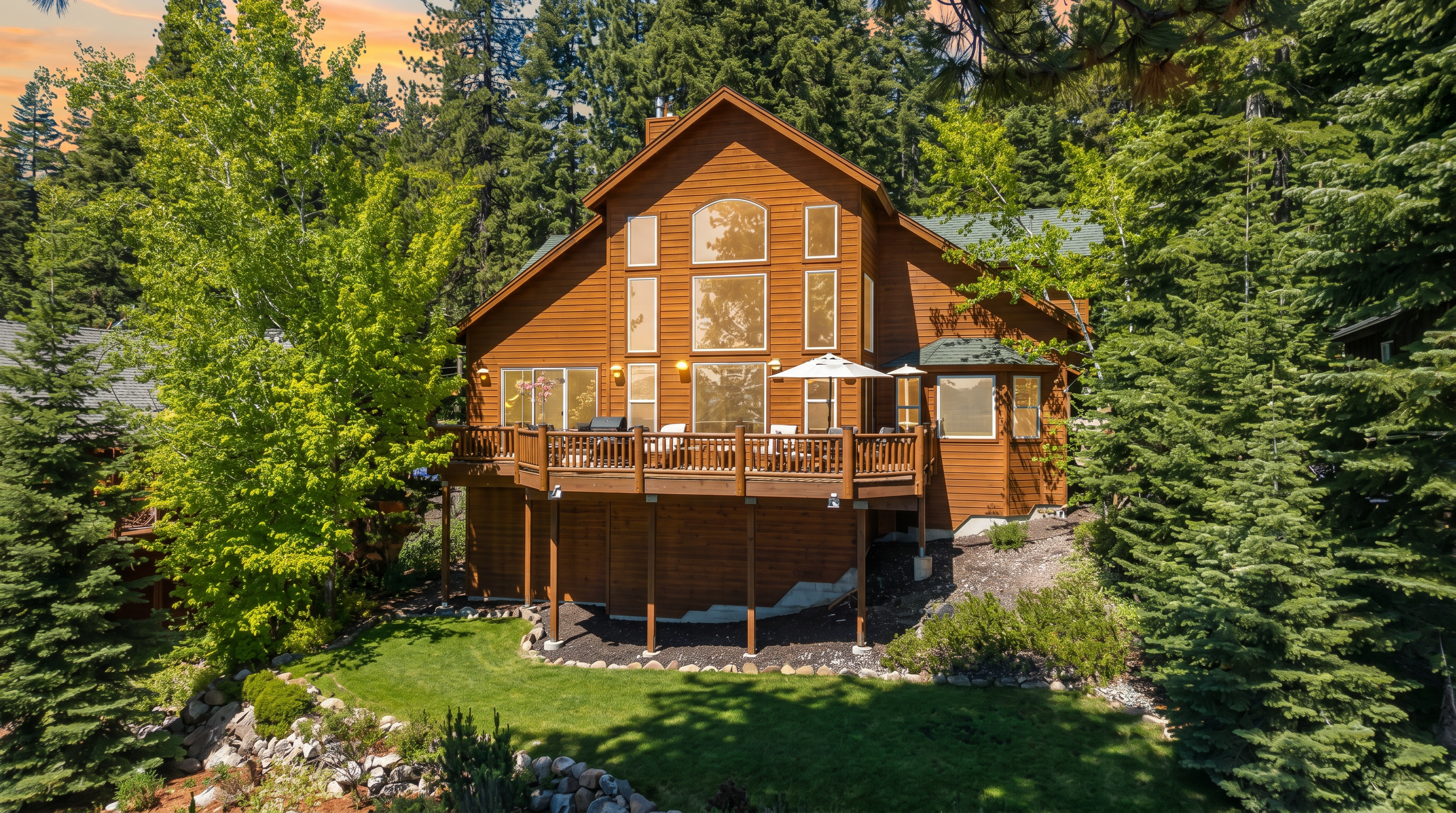 This stunning mountain cabin features a large, elevated wood deck perfect for outdoor entertaining, set against a backdrop of lush evergreen trees. The home boasts a classic rustic architectural style with wood siding and an expansive multi-story window arrangement that allows for abundant natural light. The well-manicured yard, complete with a stone-bordered lawn and dense forest surround, offers a private and serene woodland living experience.
