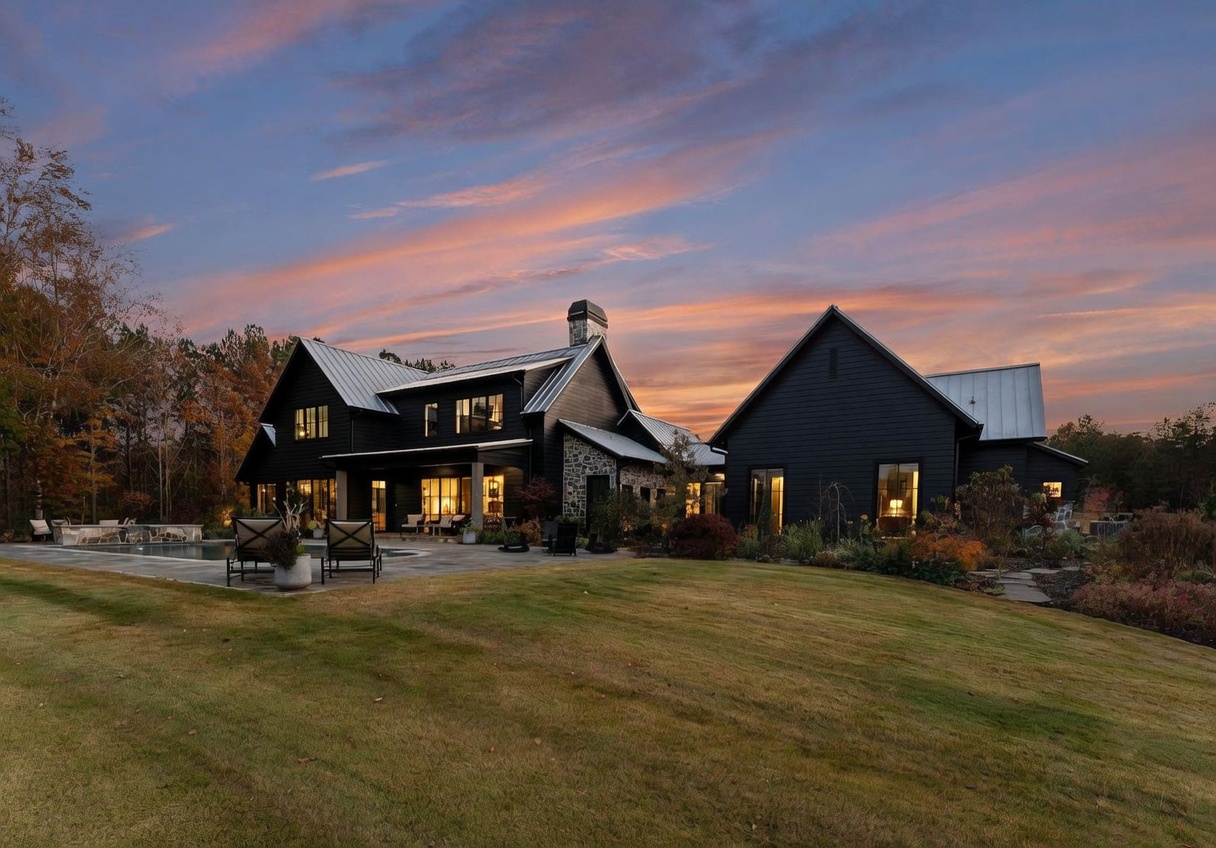 This striking modern farmhouse features dark-painted siding, a metal roof, and a prominent stone-clad chimney breast that anchors the rear facade. A spacious stone patio extends from the home, complete with outdoor seating and a swimming pool, overlooking a manicured lawn. The scene is captured at dusk, with the warm glow of interior lighting contrasting against a vibrant, colorful sunset sky.