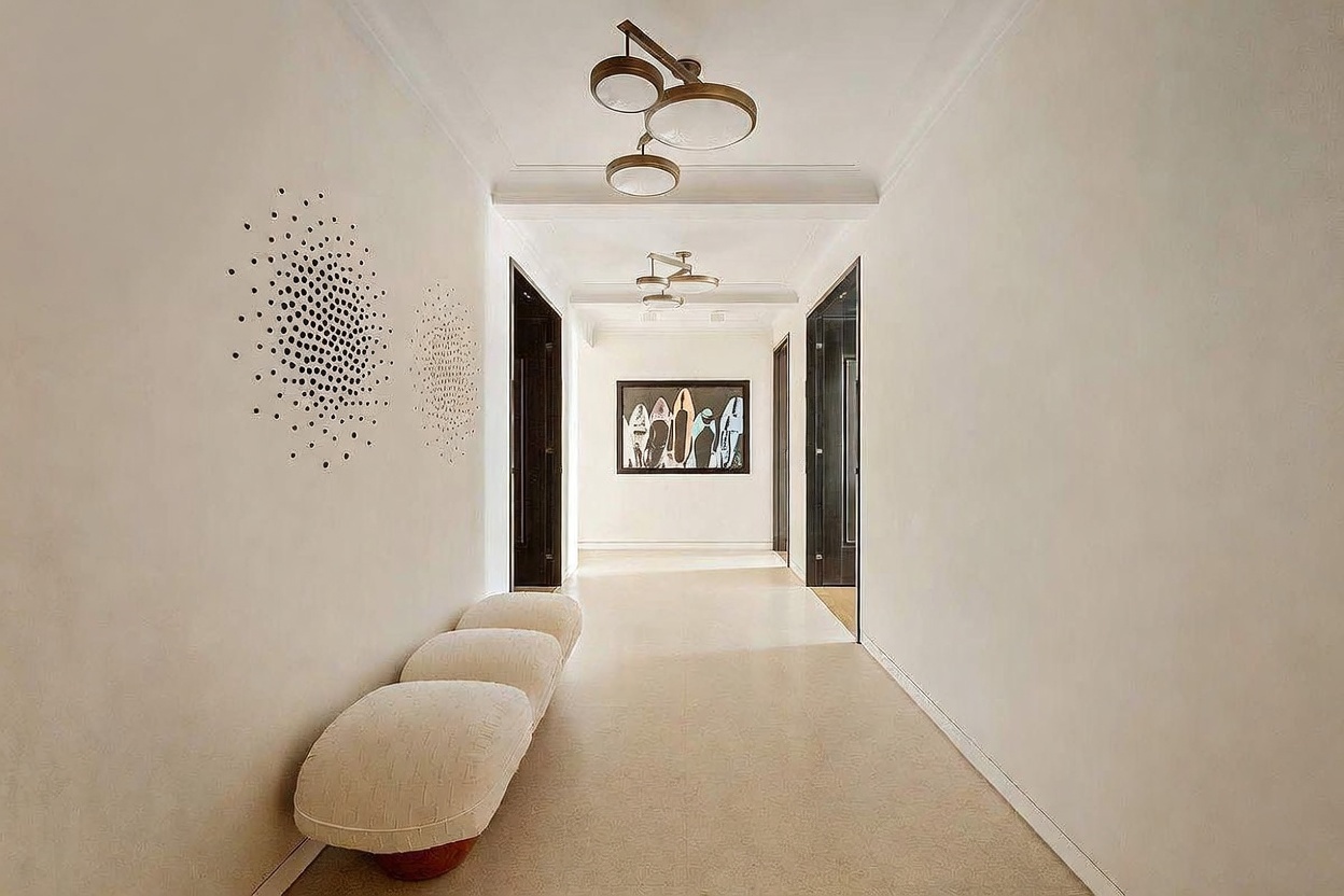 This elegant, minimalist hallway features crisp white walls and a neutral-toned carpet, creating a bright and airy atmosphere. The space is accented by a unique, sculptural light fixture and a series of textured, circular wall art pieces, while a modern bench provides a functional seating area. A framed piece of art at the end of the corridor draws the eye, emphasizing the clean lines and sophisticated design of the home.