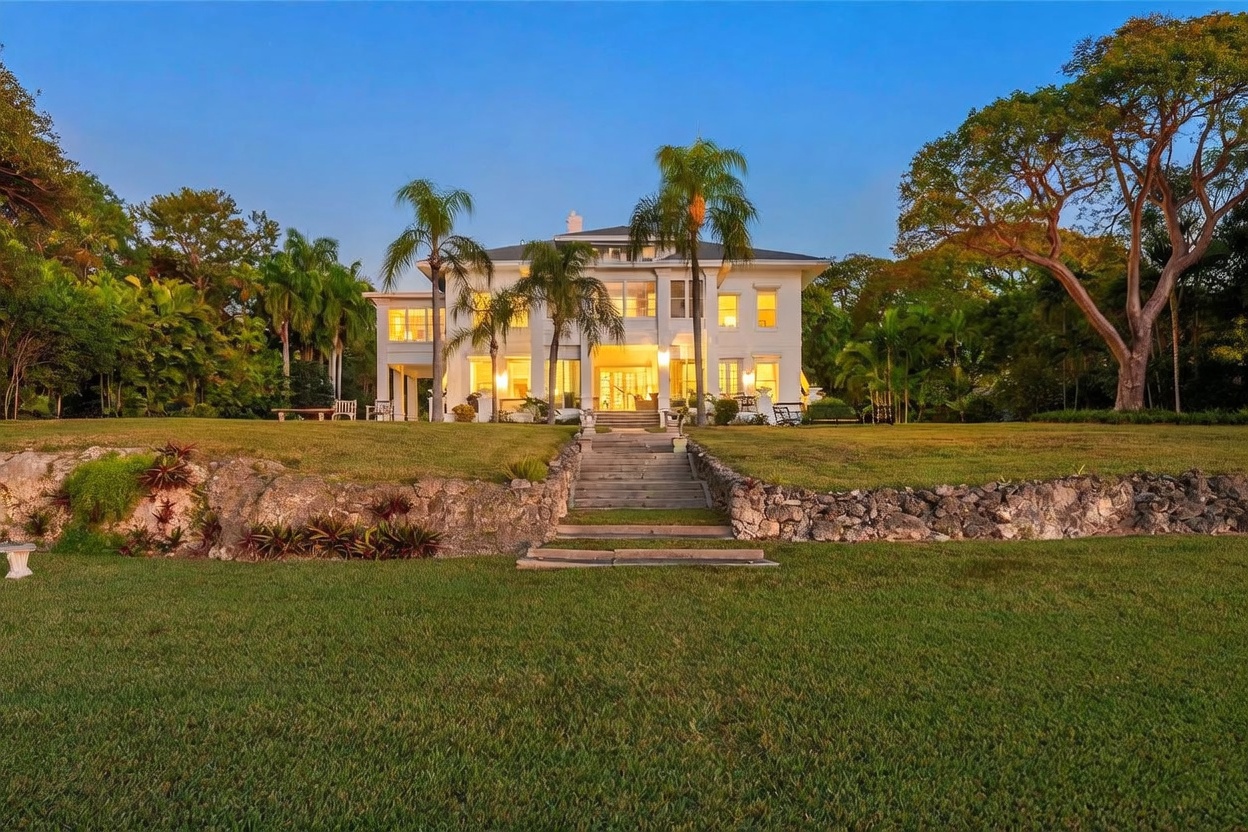 This grand, multi-story estate is captured from a low-angle perspective, emphasizing its imposing presence against a clear sky. The home features a symmetrical white facade with numerous windows, a central entryway, and a stone-retaining wall that leads to a set of stairs ascending toward the house. Lush green lawns and mature trees frame the property, creating a serene and private atmosphere.