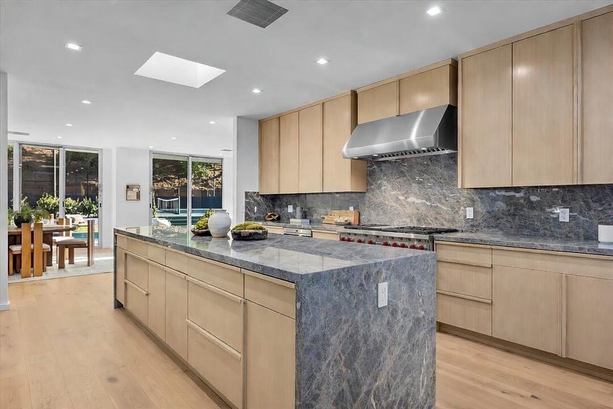 This modern kitchen features a large central island with a stone waterfall edge and light wood cabinetry. A professional-style range with a stainless steel hood is set against a dark stone backsplash, creating a sophisticated contrast. The open-concept design flows seamlessly into an adjacent dining area, with large sliding glass doors providing a view of the outdoor space.