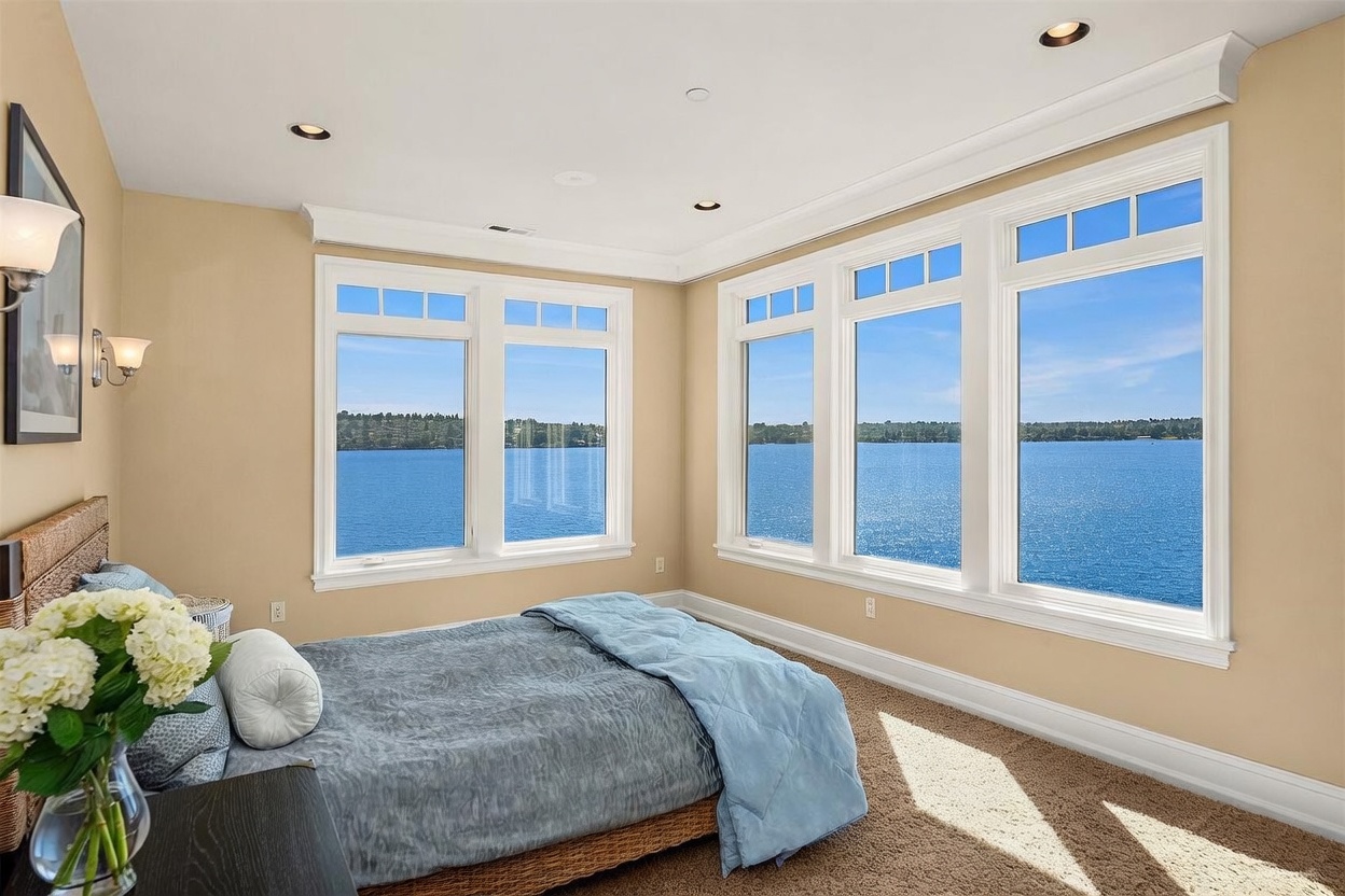 This bright and airy bedroom features a cozy bed positioned to take full advantage of the expansive, panoramic water views provided by the large windows. The room is decorated in a neutral, calming palette with soft beige walls and plush carpeting, creating a serene atmosphere. The perspective captures the seamless blend of comfortable indoor living with the tranquil beauty of the lake outside.