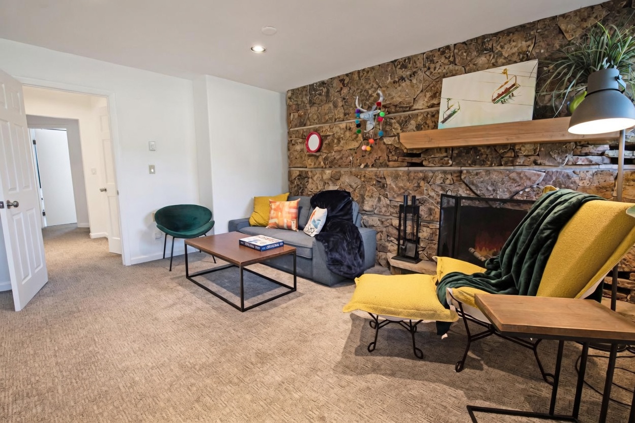 This cozy living room features a prominent stone-clad chimney breast that serves as a rustic focal point, complemented by a wooden mantel and a fireplace. The space is furnished with a grey sofa, a green accent chair, and a coffee table, all set against neutral carpeting. A doorway leads to an adjacent room, and the room is accented with colorful decor, including a whimsical wall hanging and vibrant throw pillows.